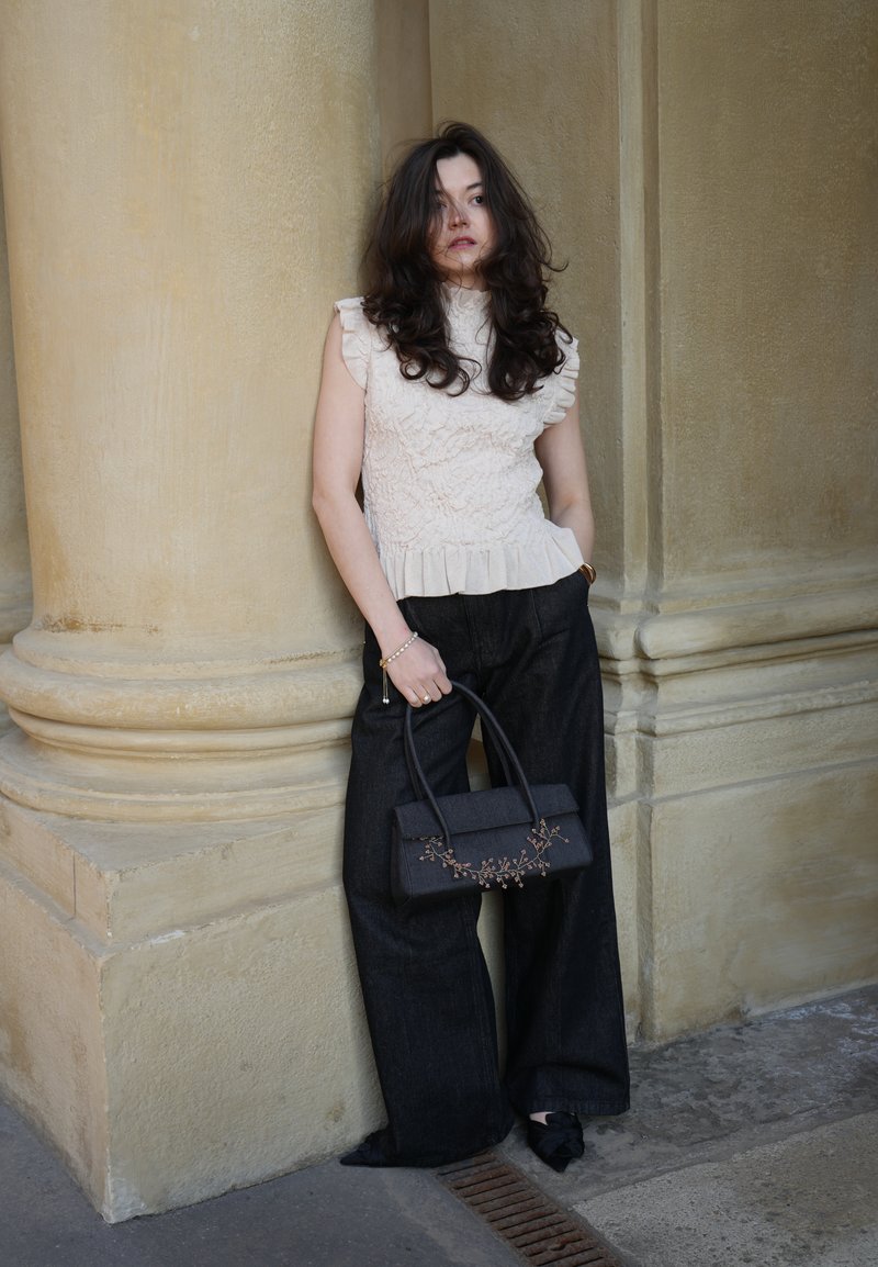 Kobieta o falowanych ciemnych włosach, ubrana w białą, bezrękawnikową bluzkę z falbanami, szerokie czarne spodnie i czarne, spiczaste buty, trzymająca czarną torebkę, opierająca się o beżową kolumnę.