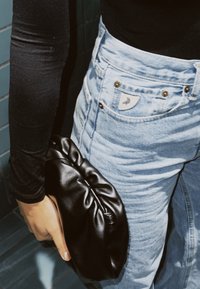 Personne portant un jean bleu clair et un haut noir à manches longues, tenant une petite pochette en cuir noir à côté d'elle.