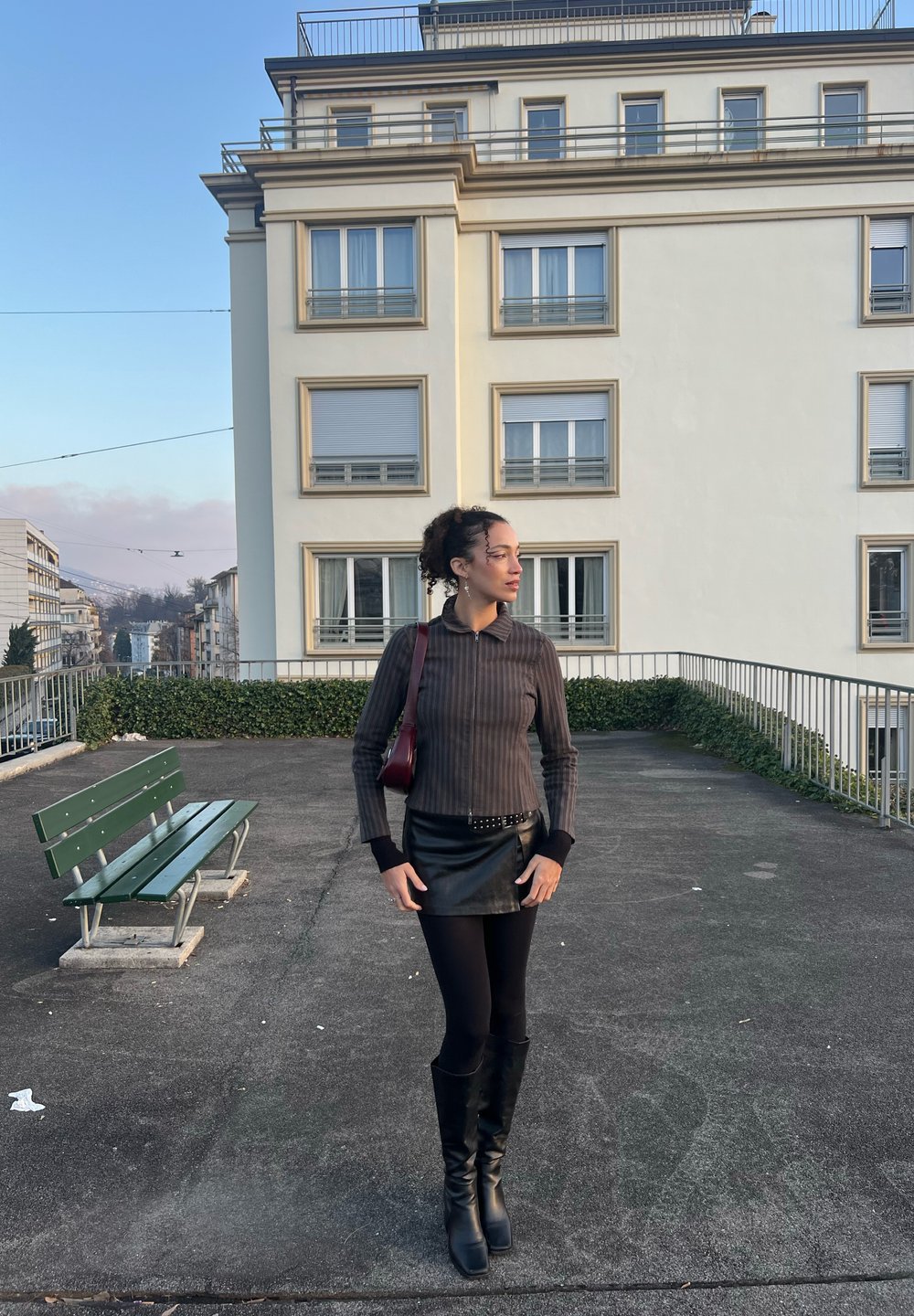 Mini-jupe en cuir noir, haut à manches longues gris foncé côtelé avec col, bottes noires montant jusqu'aux genoux et petit sac à main rouge. Fond en béton.