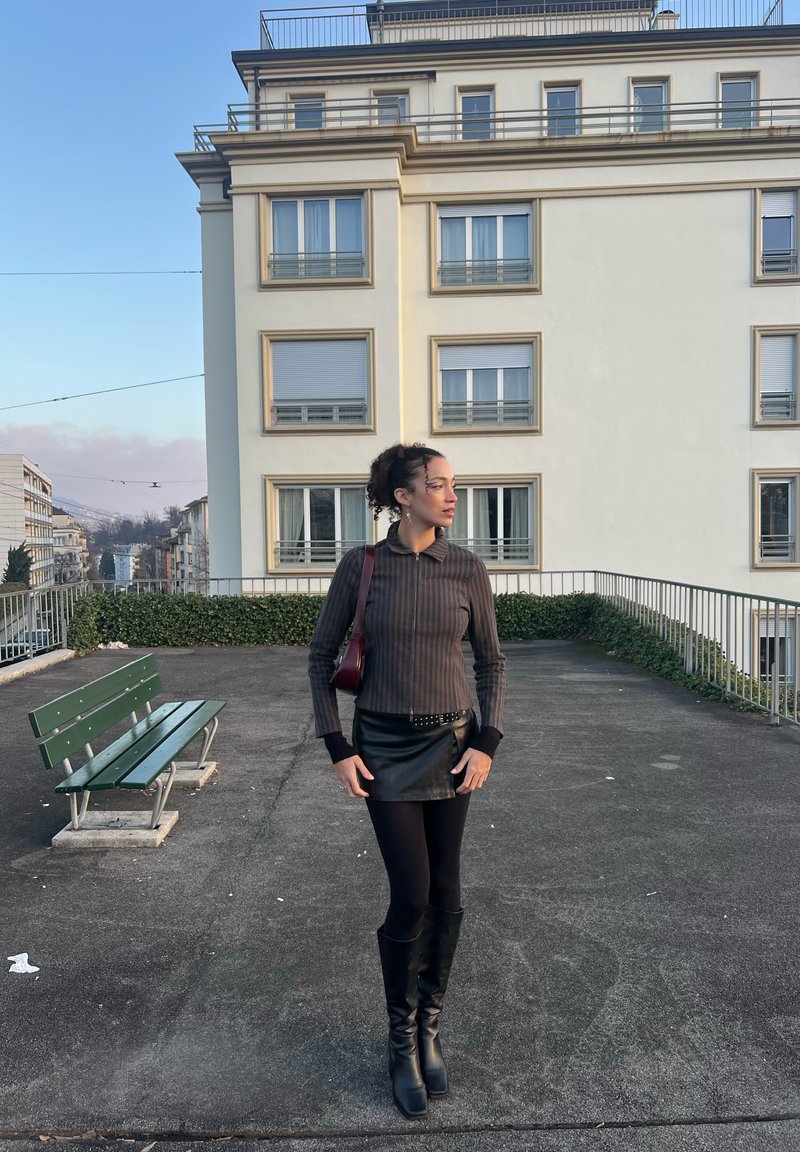 Mini-jupe en cuir noir, haut à manches longues côtelé gris foncé avec col, bottes noires montant aux genoux, et petit sac à main rouge. Fond en béton.