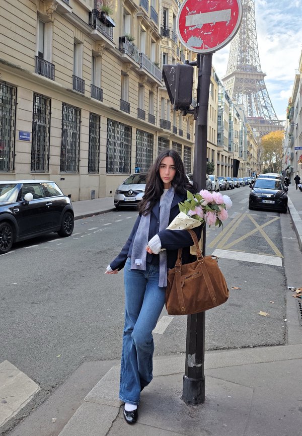 Bolso de ante marrón con un bolsillo con cremallera, vaqueros azules, calcetines blancos y una bufanda a rayas frente a una escena callejera con la Torre Eiffel al fondo.