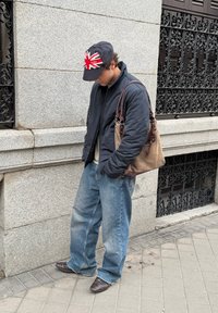 Homme portant une veste matelassée bleu marine, une chemise de couleur claire et un jean bleu ample. Tenant un sac en tissu marron avec des accents en cuir. Casquette noire avec un motif Union Jack rouge et blanc.