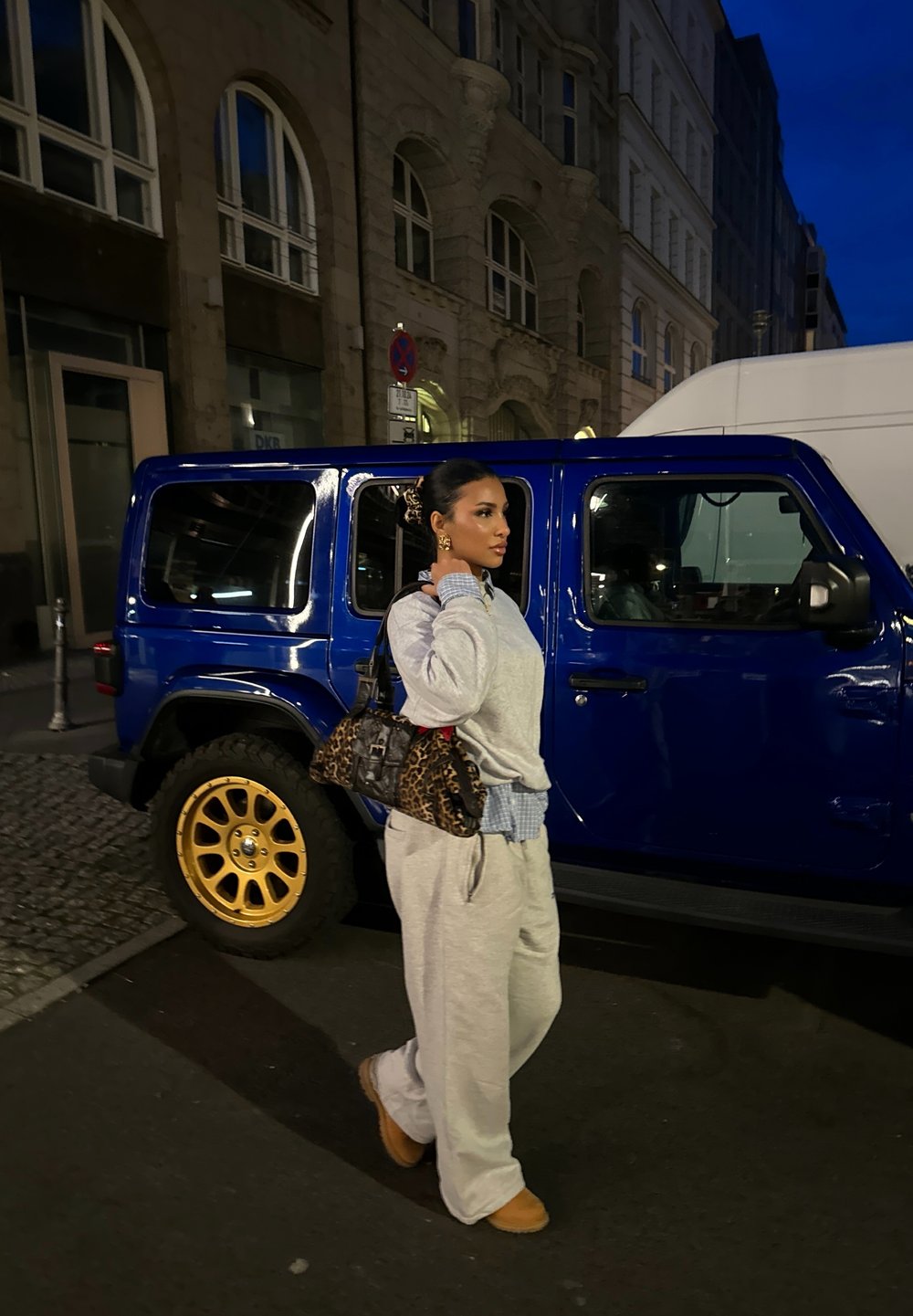 Γκρι φούτερ και φόρμες με μπλε καρό πουκάμισο από κάτω, κρατώντας μια τσάντα με λεοπάρ σχέδιο. Μπλε τζιπ με χρυσές ζάντες παρκαρισμένο κοντά.