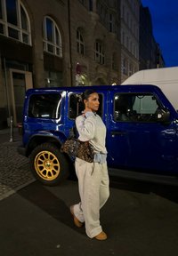 Γκρι φούτερ και φόρμες με μπλε καρό πουκάμισο από κάτω, κρατώντας μια τσάντα με λεοπάρ σχέδιο. Μπλε τζιπ με χρυσές ζάντες παρκαρισμένο κοντά.