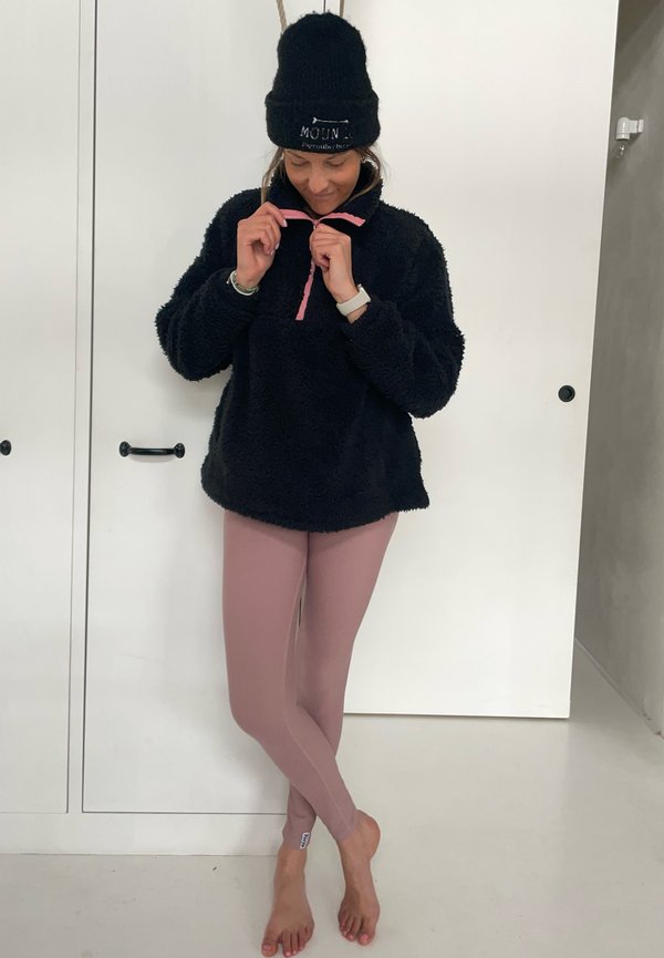 Pullover de fleece preto com meia abertura e detalhes em rosa, combinado com leggings rosa claro. Descalço, contra um fundo simples.