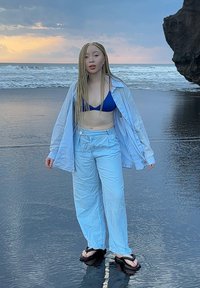 Jasnoniebieska koszula z guzikiem na wierzchu bikini w niebieskim kolorze, zestawiona z luźnymi niebieskimi spodniami i czarnymi klapkami, stojąca na mokrym piasku przy oceanie.