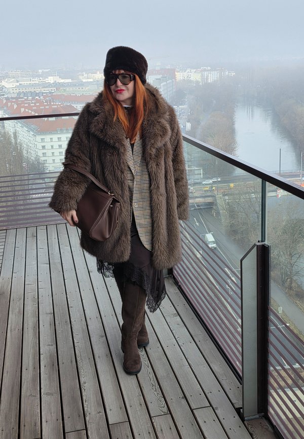 Femeie cu păr roșu, purtând o haină maro de blană, pălărie, ochelari de soare și cizme, stă pe balcon cu vedere spre oraș și râu într-o zi cețoasă.