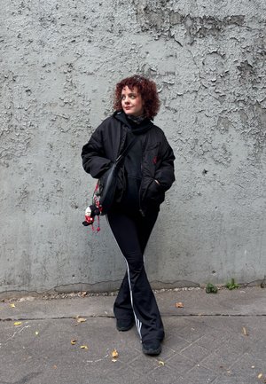 Cazadora bomber negra sobre una sudadera con capucha negra, combinada con pantalones deportivos negros con franjas blancas. Zapatos negros y un bolso bandolera con colgantes.