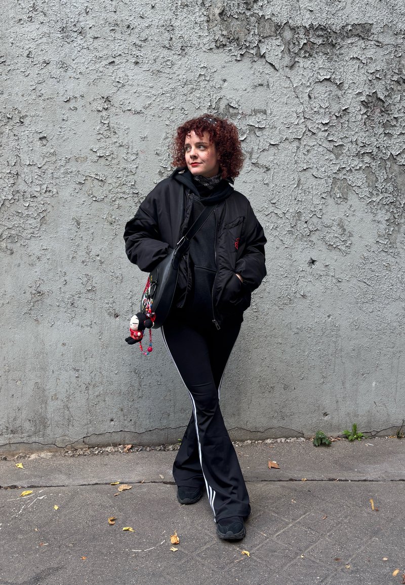 Giacca bomber nera sopra una felpa con cappuccio nera, abbinata a pantaloni sportivi neri con strisce bianche. Scarpe nere e una borsa a tracolla con ciondoli.