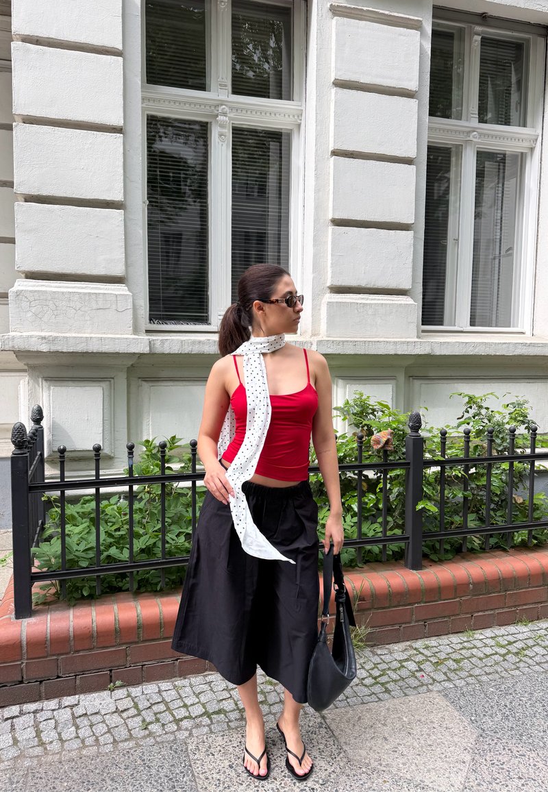 Mujer con camiseta sin mangas roja, falda negra, pañuelo blanco con lunares y sandalias, de pie en la acera de la ciudad cerca de una valla de hierro negra y un edificio blanco.