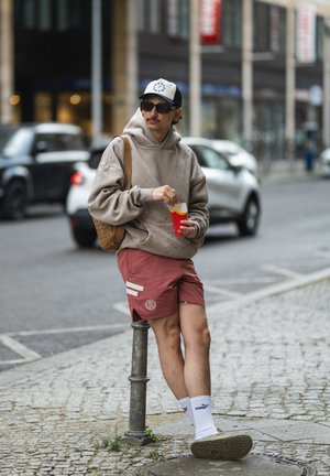Asmuo, dėvintis kreminės spalvos didelę kapšą ir raudonas šortus su baltais dryžiais, laiko raudoną puodelį ir stovi šalia metalinio stulpelio.