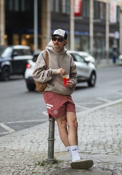 Individuo que lleva puesto un sudadera oversized beige y pantalones cortos rojos con rayas blancas, sosteniendo un vaso rojo, de pie junto a un poste de metal.