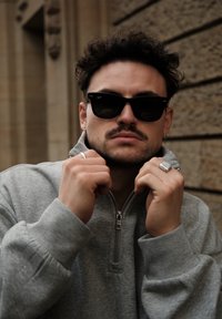 Muž s kučeravými vlasmi, ktorý nosí čierne slnečné okuliare a sivú mikinu na zips, si upravuje golier na pozadí kamennej steny.