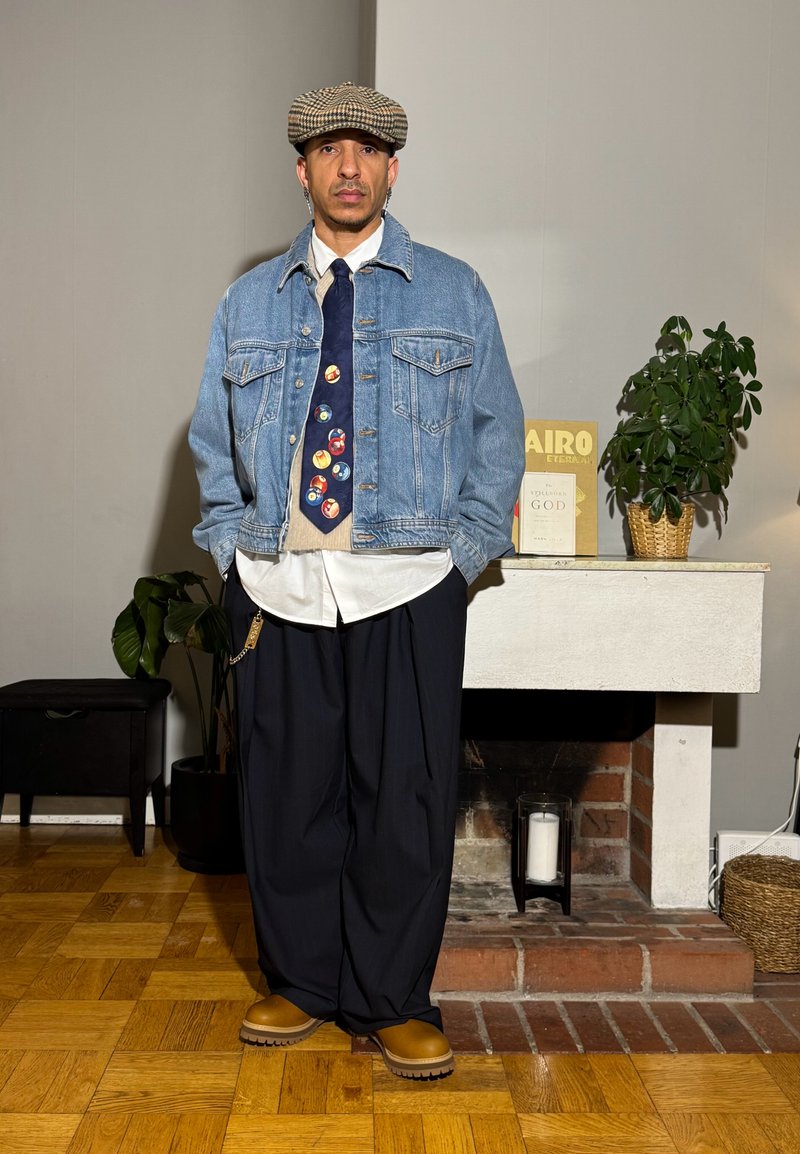 Homme portant une casquette plate à carreaux, une veste en jean, une chemise blanche, une cravate colorée, un pantalon large noir et des bottes beige, debout dans une pièce avec des plantes et des livres.