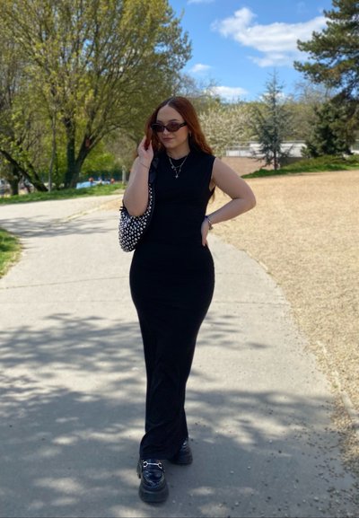 Vestido midi negro sin mangas, silueta ajustada, textura suave. Los accesorios incluyen un bolso con tachuelas y gafas de sol oscuras. Entorno casual al aire libre.