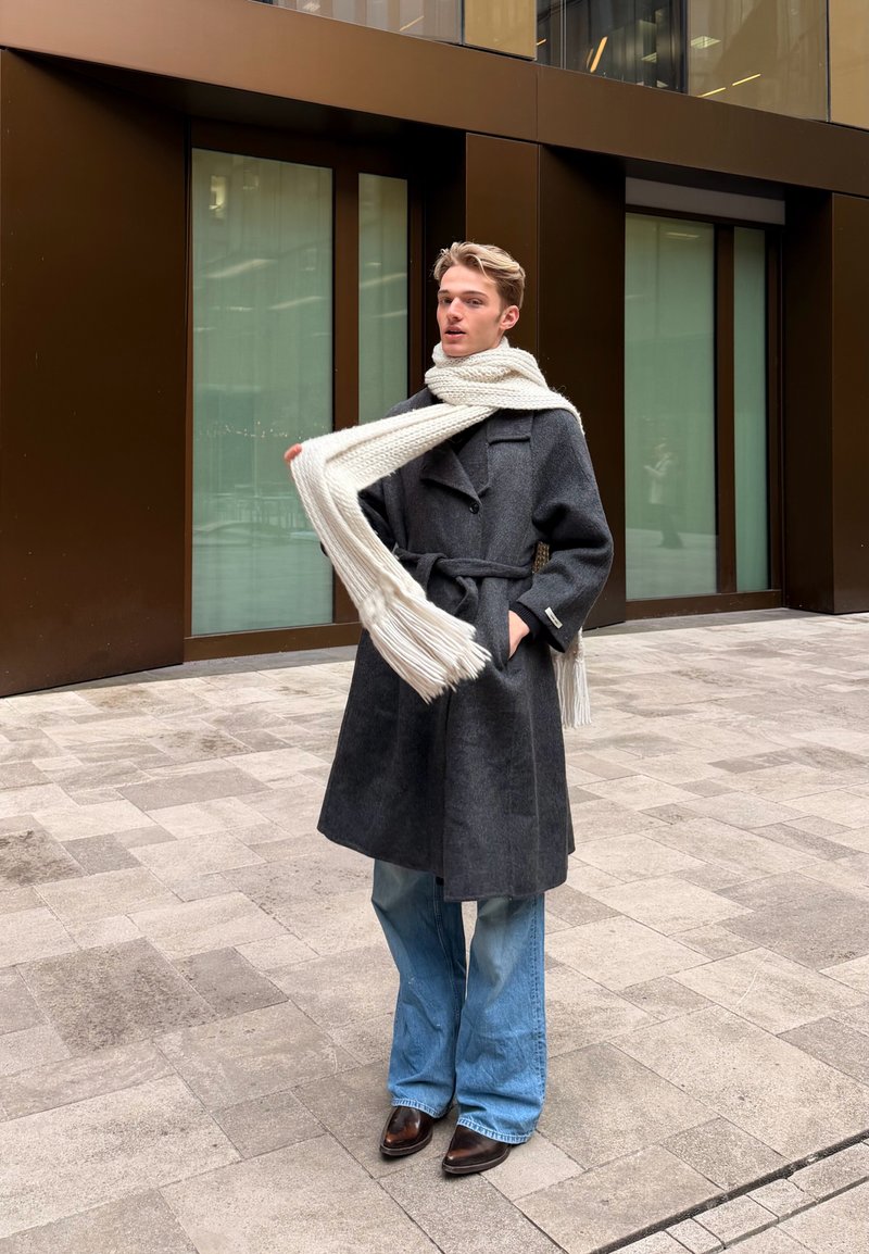 Giovane persona in piedi all'aperto con cappotto grigio con cintura, ampia sciarpa bianca, jeans svasati e scarpe marroni di fronte a un edificio moderno con grandi finestre.
