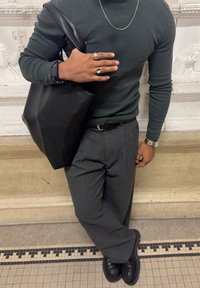 Homme en chemise à manches longues grise et pantalon gris appuyé contre un mur, portant un sac en cuir noir sur l'épaule, arborant des bagues, une montre et une chaîne en collier.