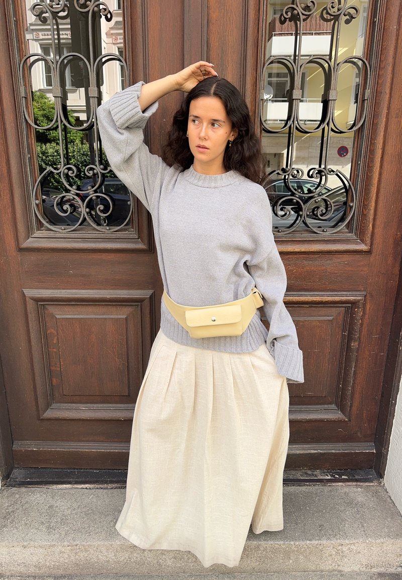 Maglione grigio chiaro lavorato a maglia con polsini a costine, abbinato a una gonna beige a vita alta e plissettata e a una cintura gialla, davanti a una porta in legno.