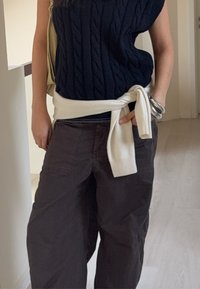 Pull en maille torsadée bleu marine, pull blanc nou é à la taille, pantalon ample foncé, bracelets argentés au poignet, décor intérieur minimaliste.