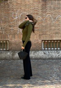 Pulover verde măsliniu cu dungi, cu guler din blană, asortat cu pantaloni evazați negri și o geantă mică neagră. Fundalul prezintă un perete de cărămidă.