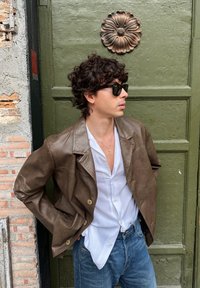 Veste en cuir marron avec une coupe légèrement oversize, chemise blanche boutonnée, et jean bleu. Accessoires : lunettes de soleil noires. Fond mur vert.