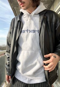 Jaqueta de couro preta sobre um hoodie cinza claro com o logo "carhartt", combinada com uma camisa branca e calças pretas risca de giz. Materiais texturizados visíveis.