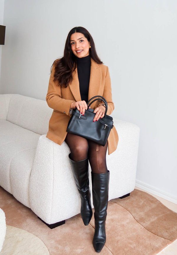 Sieviete sēž uz baltas dīvāna, valkājot kamieļa krāsas žaketi, melnu augstu apkakli, melnu svārku, ceļgalu augstus zābakus, turot melnu ādas rokassomu.