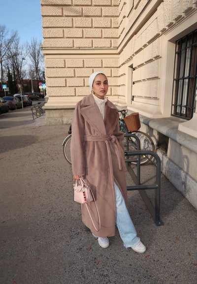 Abrigo de pelo sintético marrón claro con un cinturón en la cintura, llevado sobre un suéter de cuello alto crema, combinado con jeans azul claro y un pequeño bolso rosa.