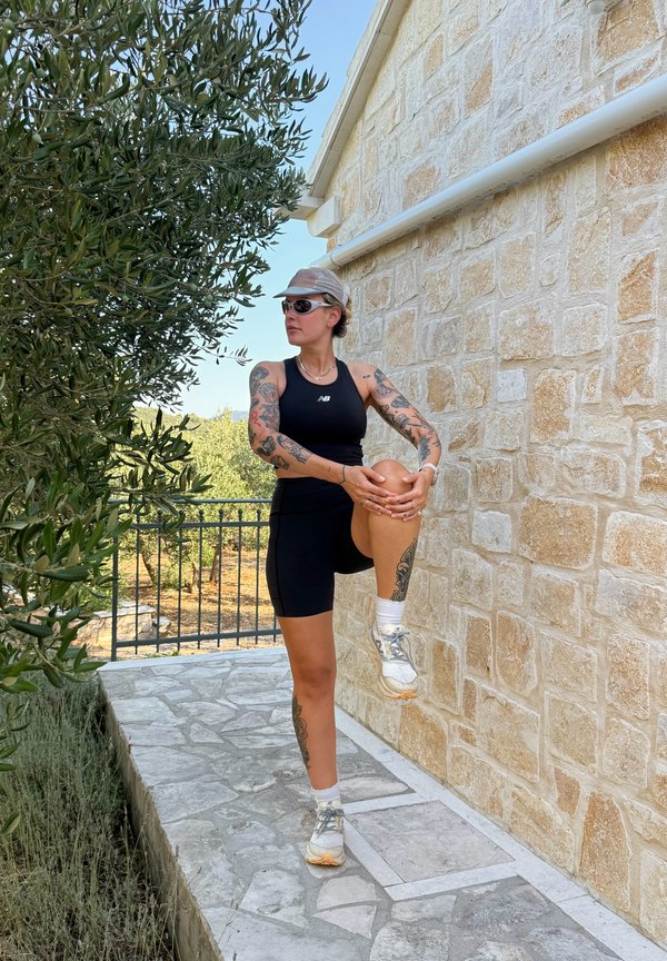 Mujer con tatuajes en ropa deportiva estirando su pierna levantada cerca de una pared de piedra afuera en un día soleado.