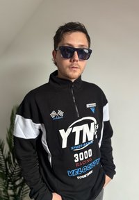 Svartvit zip-up jacka med "YTN"-märke, blå detaljer och racinginspirerade grafik. Modell bär solglasögon, poserar mot en enkel bakgrund.