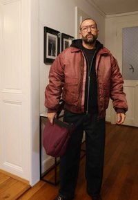 Sarkana satīna bumbvedējs jaka ar rāvējslēdzēja aizdari, divām priekšējām kabatām, uzvilkta pār melnu kapuci. Tumši džinsi un dziļi bordo krāsas soma. Koka grīda.