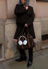 Brun fløyelshåndveske med flere glidelåslommer, hvite hodetelefoner festet, brukt med en svart oversized blazer og høye svarte støvler.