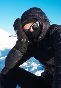 Person iført sort vinterudstyr, solbriller og vanter, der hviler hovedet på hånden i et snedækket udendørs miljø under en klar blå himmel.