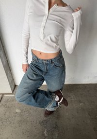 Personne portant un haut blanc à manches longues court, un jean bleu et des chaussures marron, debout sur une jambe contre un mur blanc.