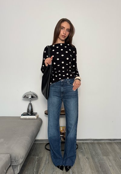 Mujer con blusa negra de manga larga con lunares y jeans azules, de pie en el interior sosteniendo un bolso negro de hombro, junto a una mesa con libros y una lámpara.