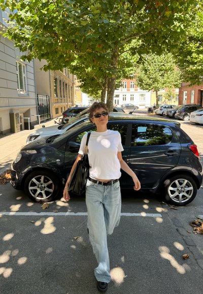 Bolso negro, camiseta corta blanca, jeans de pierna ancha desvanecidos y botas negras gruesas. Auto negro estacionado y árboles frondosos de fondo.