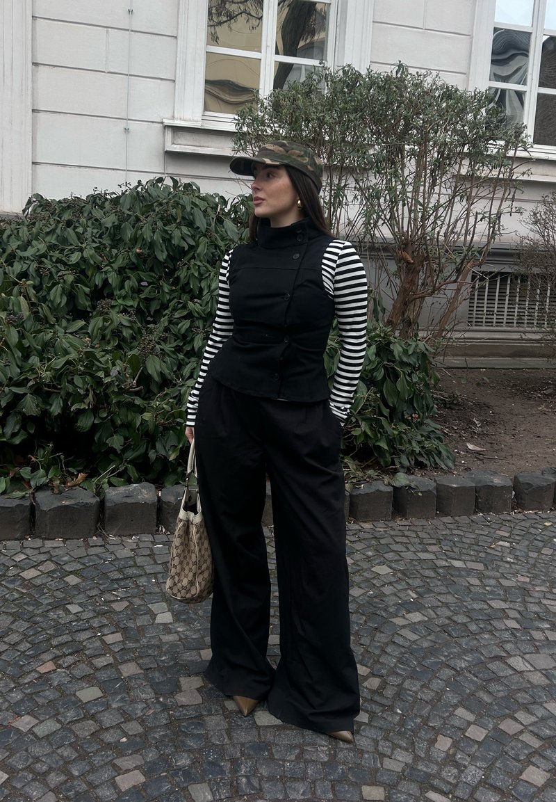 Γυναίκα που στέκεται σε λιθόστρωτο δρόμο κοντά σε θάμνους και κτίριο, φορώντας ριγέ μπλούζα με μακριά μανίκια, μαύρο γιλέκο, φαρδιά παντελόνια, καπέλο παραλλαγής, κρατώντας τσάντα με μοτίβο.