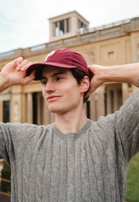 Casquette bordeaux avec une lettre à l'avant, pull gris tricoté avec un motif torsad é, posé en extérieur devant des structures architecturales.