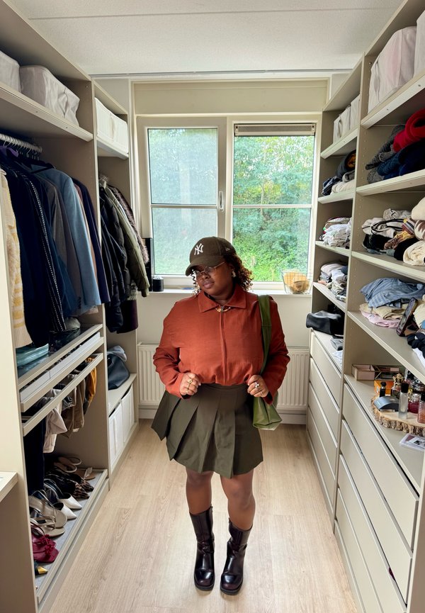 Personne portant une veste marron, une jupe verte, des bottes noires et une casquette de baseball, debout dans un dressing avec des étagères et des vêtements suspendus.