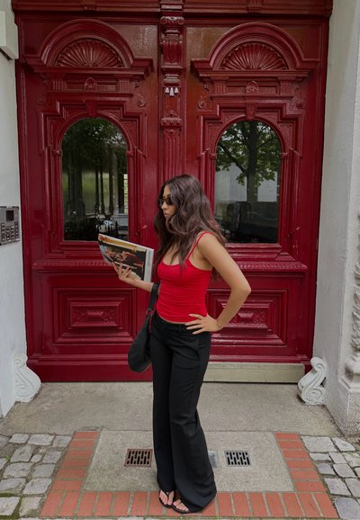 Mujer con cabello largo y oscuro y gafas de sol está delante de unas grandes puertas dobles rojas y ornamentadas, sosteniendo una revista y vistiendo una blusa roja y pantalones negros.
