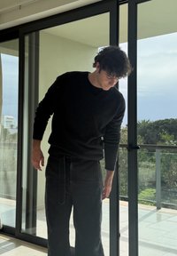 Νεαρός άνδρας με μαύρο πουλόβερ και παντελόνι σκύβει μπροστά κοντά σε γυάλινη πόρτα με μπαλκόνι και ορατό πράσινο έξω.