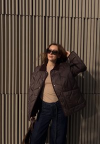 Brun quiltet pufferjakke med mørk krage, båret over en beige topp og mørke jeans. Solbriller og en brun veske er også til stede.