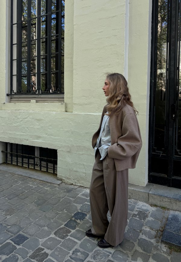 Vrouw met lang golvend haar in een beige jas en wijde broek staat op een kasseienstraat naast een bleekgeel gebouw met grote ramen en een zwarte deur.