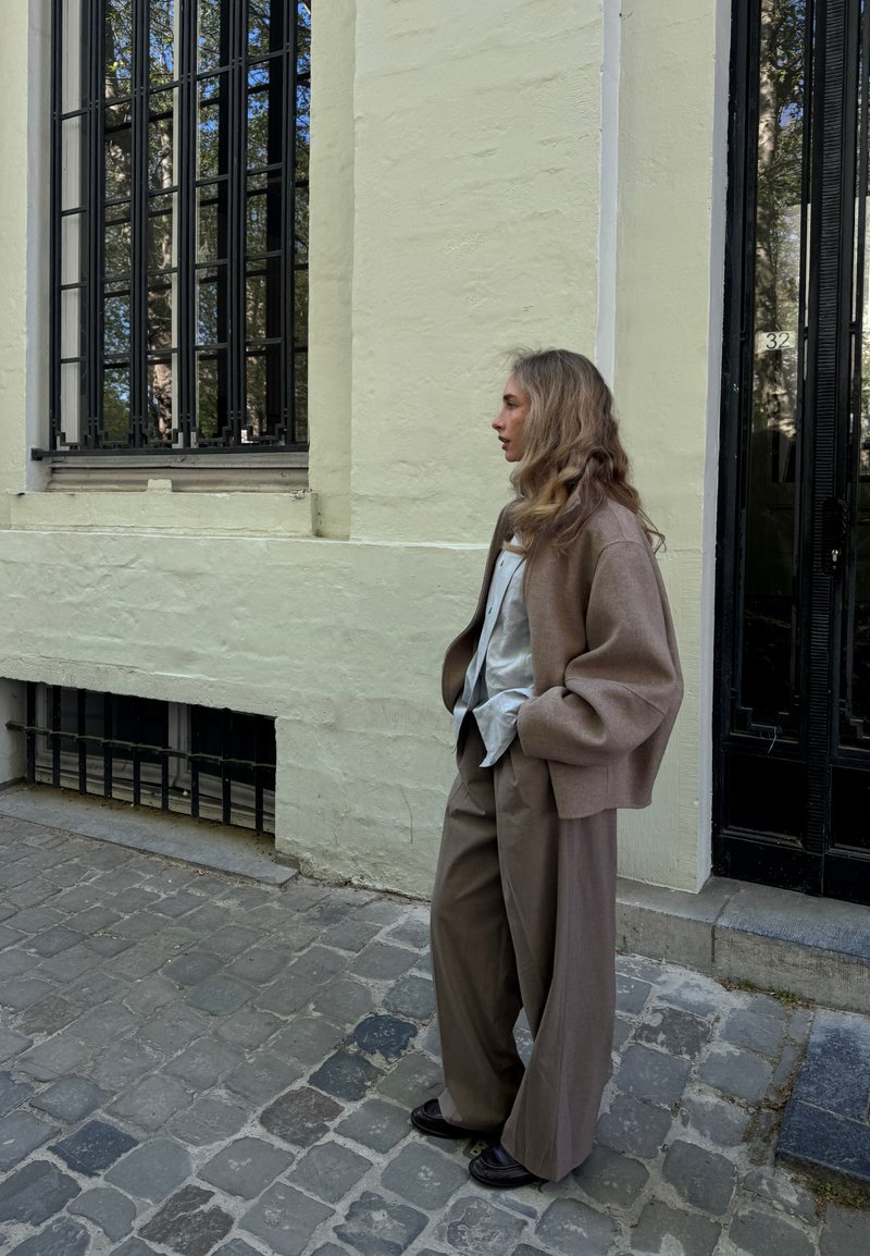 Γυναίκα με μακριά κυματιστά μαλλιά, με μπεζ σακάκι και φαρδιά παντελόνια, στέκεται σε έναν λιθόστρωτο δρόμο δίπλα σε ένα απαλό κίτρινο κτίριο με μεγάλα παράθυρα και μαύρη πόρτα.