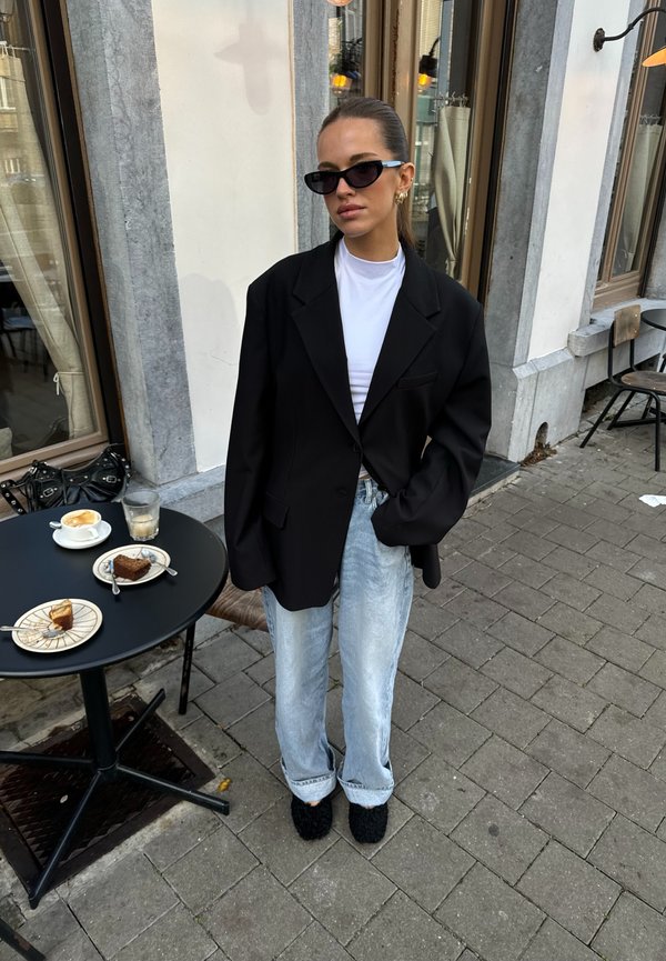 Černý oversized blejzr přes bílý triko, světle modré široké džíny a černé chlupaté pantofle, v pozadí kavárenská scenérie.