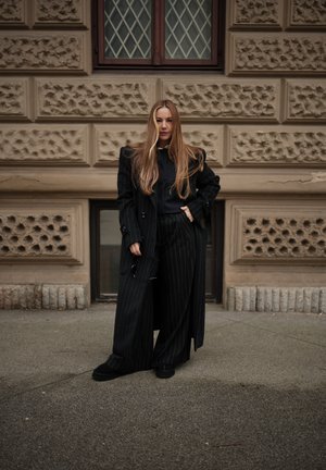 Mujer con cabello largo que lleva un abrigo negro de rayas y pantalones anchos a juego, de pie en una acera frente a un edificio de piedra con textura.