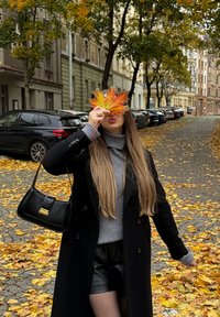 Žena v černém kabátu a šedém svetru drží velký podzimní list před obličejem na ulici pokryté listím s odstavenými automobily.