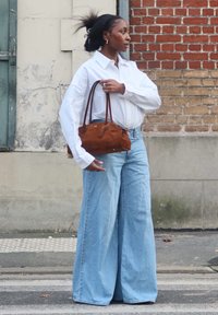 Biała, oversize'owa koszula, jasnoniebieskie dżinsy o szerokich nogawkach oraz brązowa zamszowa torebka z krótkimi uchwytami. Teksturowana ściana i ceglana ściana w tle.