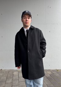 Manteau noir en laine avec un col et deux boutons, porté sur une chemise rayée et une cravate. Un jean bleu et une casquette de baseball marine complètent le look.