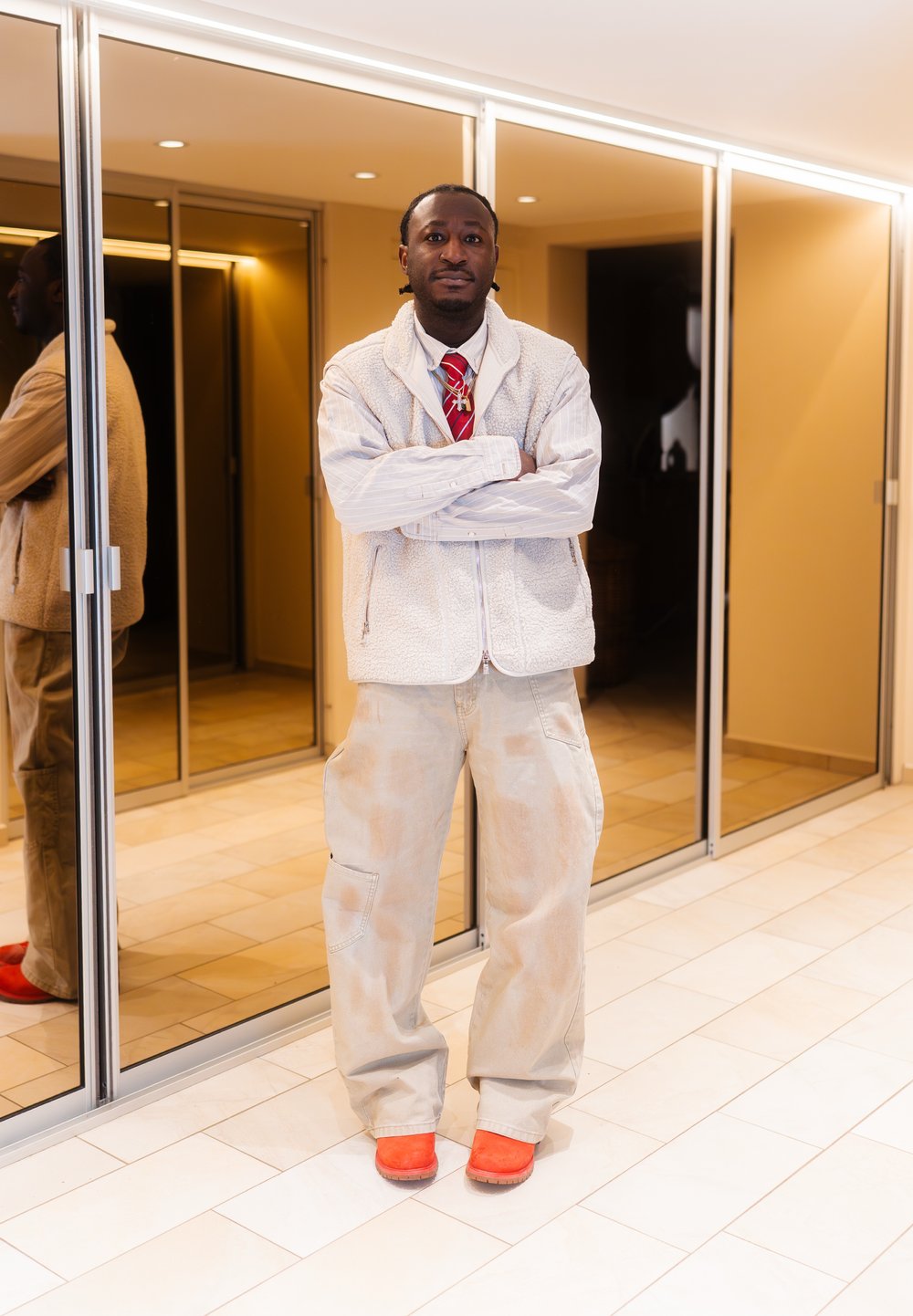 Άνδρας στέκεται με σταυρωμένα τα χέρια, φοράει λευκό γιλέκο από φλίς, ριγέ πουκάμισο, κόκκινη γρ αβάτα, μπεζ παντελόνι και φωτεινά πορτοκαλί παπούτσια σε ένα μοντέρνο διάδρομο.